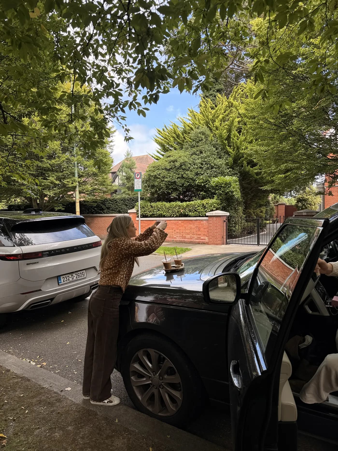 Rebeccah leaning over a car bonnet to photograph two iced coffees on a leafy Dublin street